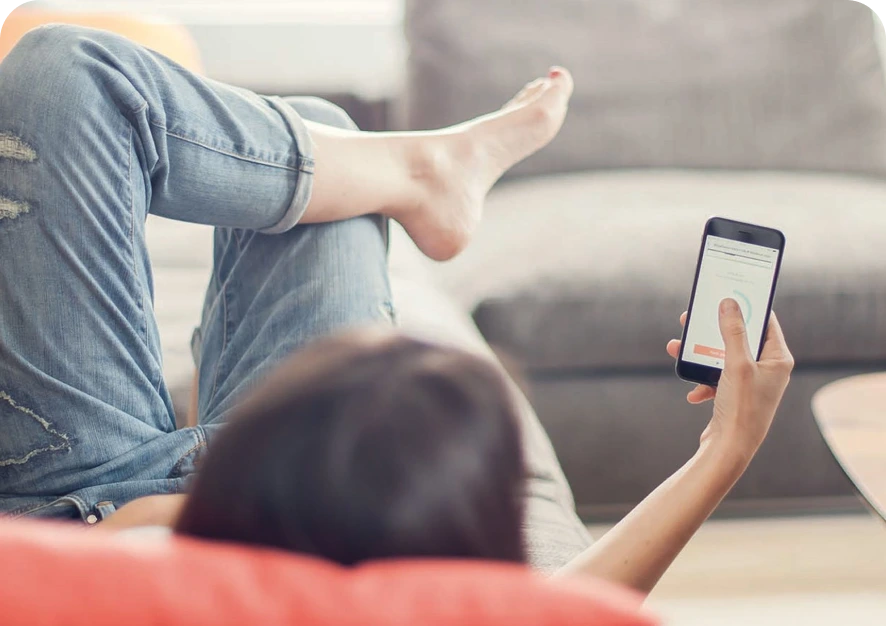 An over-the-shoulder shot of a woman lying on the couch with a smartphone in her hand. Elvie Trainer app on a phone screen.
