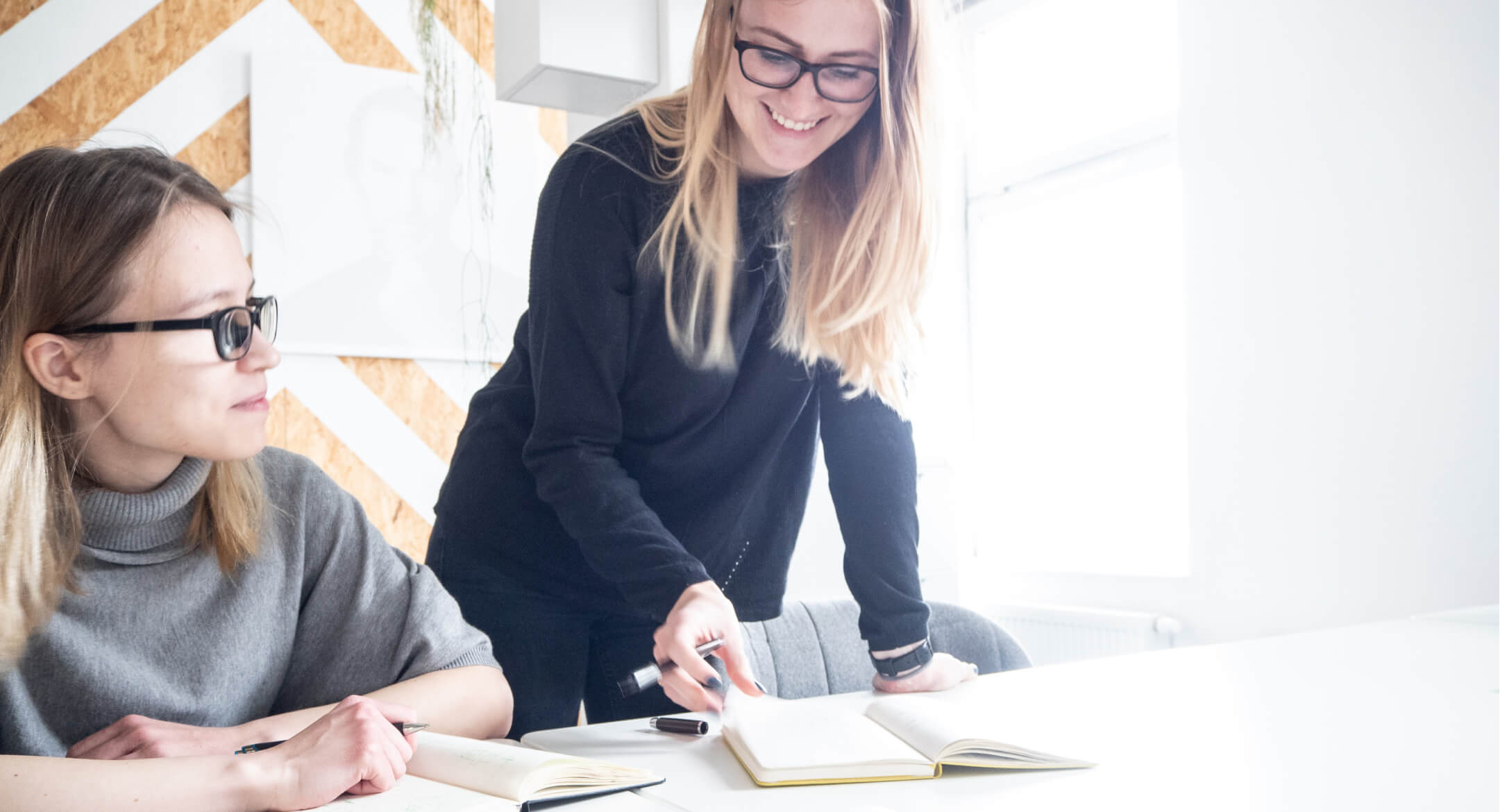 Two white women in their 20s in the Untitled Kingdom office. One is sitting, notepad on the desk. The other is standing, pointing at the notepad. Both are smiling.
