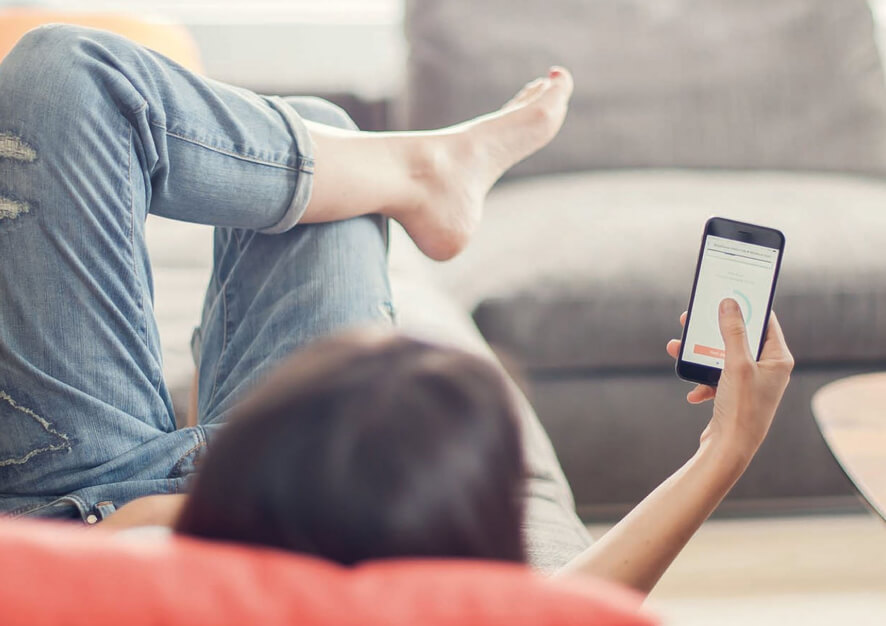 An over-the-shoulder shot of a woman lying on the couch with a smartphone in her hand. Elvie Trainer app on a phone screen.