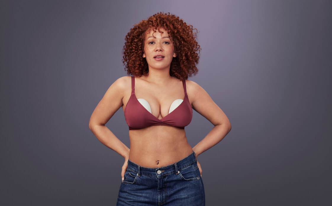 A woman standing against a grey background. She's wearing jeans and a bra with two Elvie Pumps inside. She's standing proudly with hands on her hips.