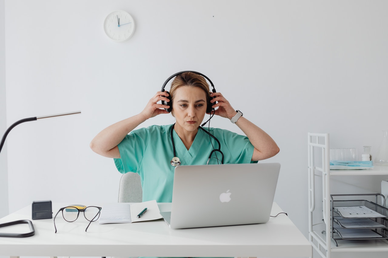 A woman in a doctor's apron sitting an office - sitting by a laptop and putting headphones on.
