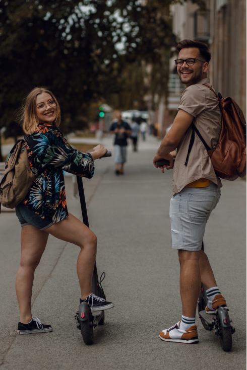 Two young people on a city street. Both are standing and smiling while leading their electric scooters.