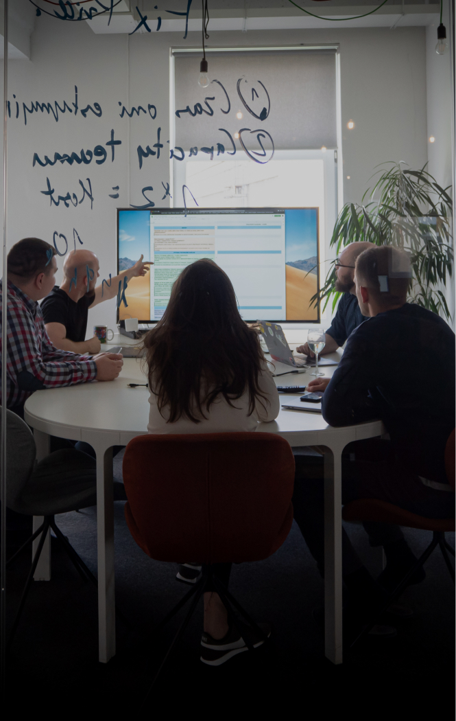 Four people sitting by the desk looking at a big screen. One person is pointing at the screen, clearly explaining something to the rest of the group.